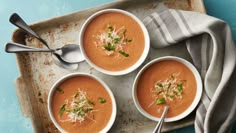 three bowls of soup on a tray with silver spoons and two napkins next to them