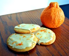 three orange cookies with icing next to an orange on a wooden table, and a bag