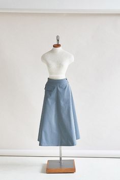 a mannequin wearing a blue skirt and white shirt on a wooden stand in front of a wall