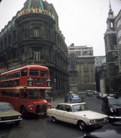 a red double decker bus driving down a street next to tall buildings and parked cars