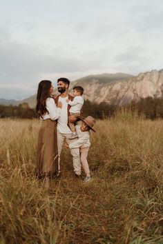 a family is standing in the tall grass with mountains behind them and one man holding two children