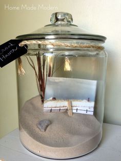 a glass jar filled with sand and some small pictures on top of the lid, sitting on a shelf