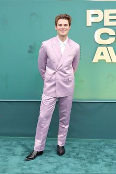 a man in a pink suit and black shoes standing on a green carpet with his hands in his pockets