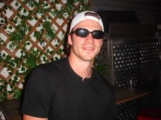 a man wearing sunglasses and a hat in front of a wall covered with ivys