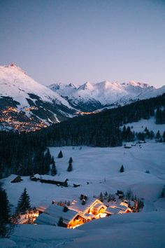 a snowy mountain with houses and lights in the foreground at night, surrounded by snow - capped mountains