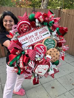 a woman standing next to a christmas wreath with cookies and milk for santa written on it