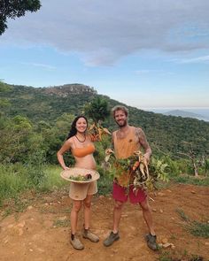 a man and woman standing next to each other on top of a dirt hill with trees