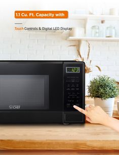 a woman pointing at a black microwave on top of a wooden kitchen counter with plants in the background