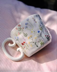 a white coffee cup sitting on top of a pink and white striped table cloth with flowers painted on it