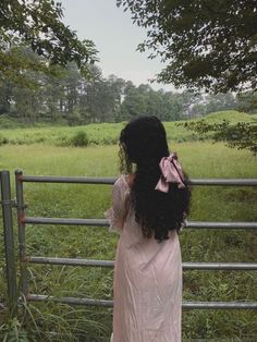 a woman standing in front of a fence looking at the grass and trees behind her