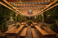 an outdoor dining area with wooden benches and lights hanging from the ceiling over it, surrounded by greenery