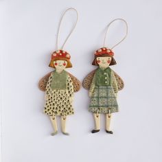 two wooden dolls are hanging from strings on a white surface, one is wearing a green dress and the other has a red hat