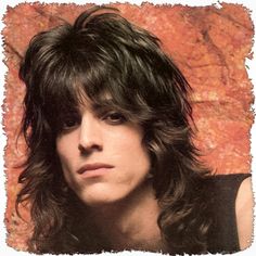 a man with long hair and black shirt posing in front of a rock wall holding a guitar