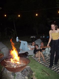 a woman standing in front of a fire pit with people sitting around it at night