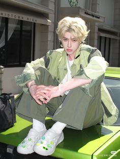 a young man sitting on the hood of a green car in front of a building