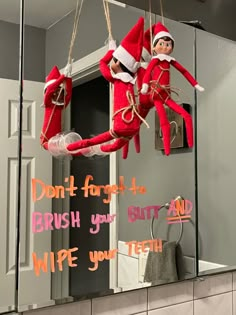 two elfs hanging from a mirror with words written on the wall above them that read don't forget to brush your teeth wipe your teeth