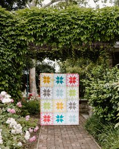 a quilt is hanging on the side of a brick walkway in front of flowers and greenery