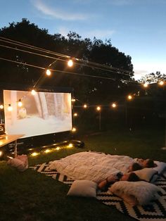 an outdoor movie is set up in the backyard with lights strung around it and people laying on blankets