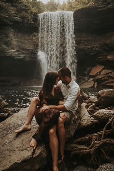 a man and woman sitting on rocks in front of a waterfall with their arms around each other