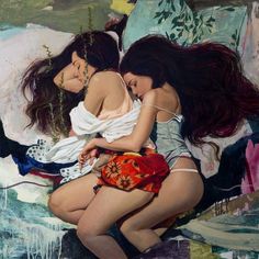 two women hugging each other while sitting on top of a bed in front of a painting