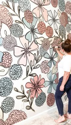 a woman standing in front of a wall with flowers painted on it and another person painting the wall behind her