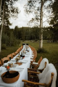 a long table is set up outside in the woods for an outdoor dinner with white linens and wooden chairs