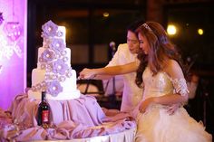 two women in wedding dresses are cutting into a white cake with purple flowers on it