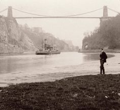 a man standing in front of a body of water near a bridge with a boat on it