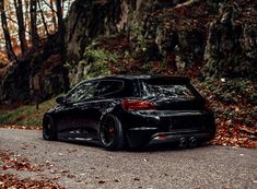 a black car parked on the side of a road in front of trees and leaves