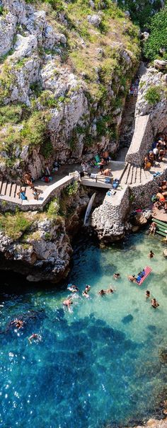 people are swimming in the clear blue water near a rocky cliff and walkway that leads to a bridge