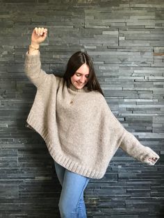 a woman standing in front of a brick wall wearing a gray sweater and blue jeans