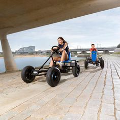 two people are riding three wheeled vehicles under a bridge over the water, with one person behind them