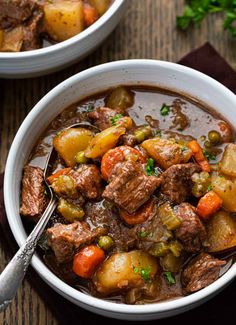 two white bowls filled with beef stew and potatoes on top of a wooden table next to a spoon