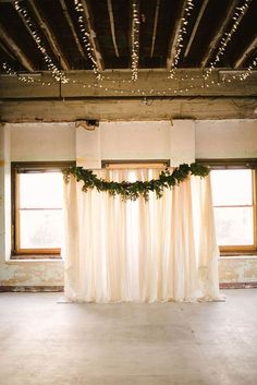 an empty room with white drapes and lights hanging from the ceiling, decorated with greenery