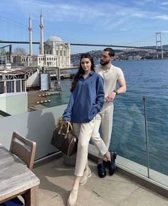 a man and woman standing on a balcony next to the water with a bridge in the background