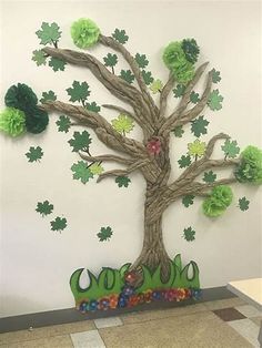 a tree made out of paper with green leaves and flowers on the branches, sitting in front of a white wall