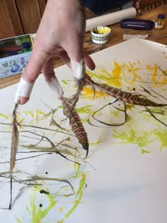 a person is painting some branches with yellow and green paint on the paper while another hand holds something in front of them