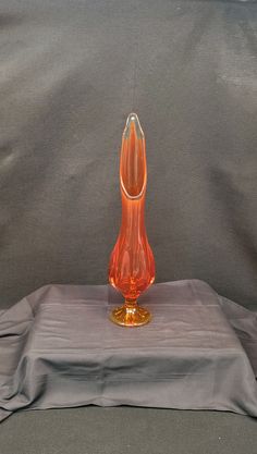 an orange glass vase sitting on top of a gray cloth covered table with a grey background