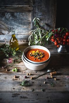 a bowl of tomato soup on a wooden table next to some flowers and olives