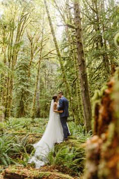 a bride and groom standing in the middle of a lush green forest with tall trees