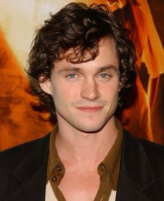 a man with curly hair wearing a black suit and brown shirt is looking at the camera