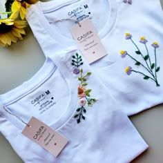 two white t - shirts with embroidered flowers on them and tags attached to the front