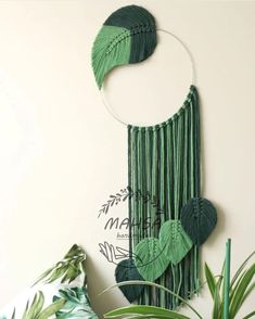 a green wall hanging with leaves on it and a plant in the corner next to it