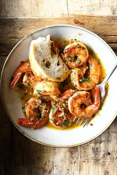 a white bowl filled with shrimp and bread on top of a wooden table next to a fork