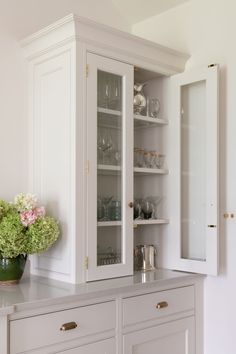 a white cabinet with glass doors and flowers in the vase next to it on top of a counter