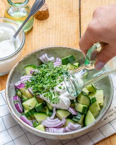 a person pouring dressing into a bowl filled with cucumber, onions and other vegetables