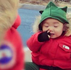 a toddler in a red coat is talking on the phone and wearing a green hat