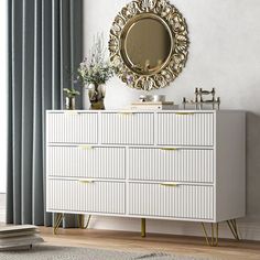 a white dresser with gold accents and a round mirror above it, in front of a window