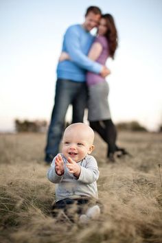 a baby sitting in the middle of a field with his mother and father behind him