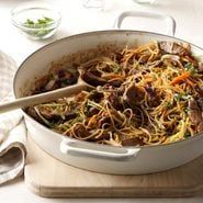 a pan filled with pasta and vegetables on top of a cutting board next to a wooden spoon